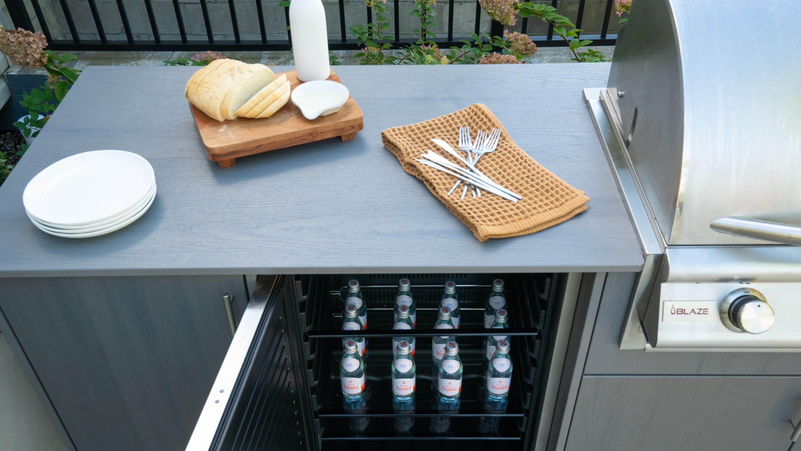Outdoor kitchen countertop with bread, drinks in under-counter refrigerator, and modern stainless grill.